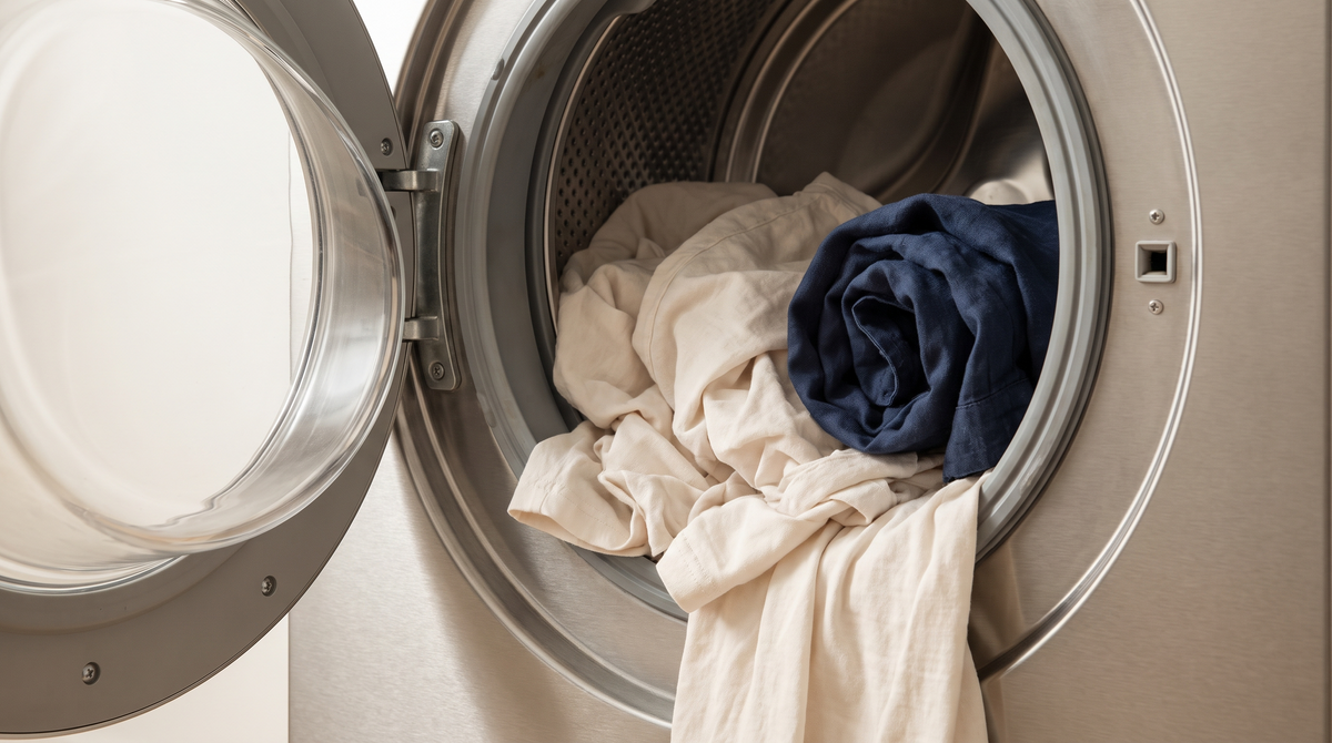 Pale cotton garments and a navy linen shirt left loose inside a brushed steel washing machine drum seen through the open door in soft daylight