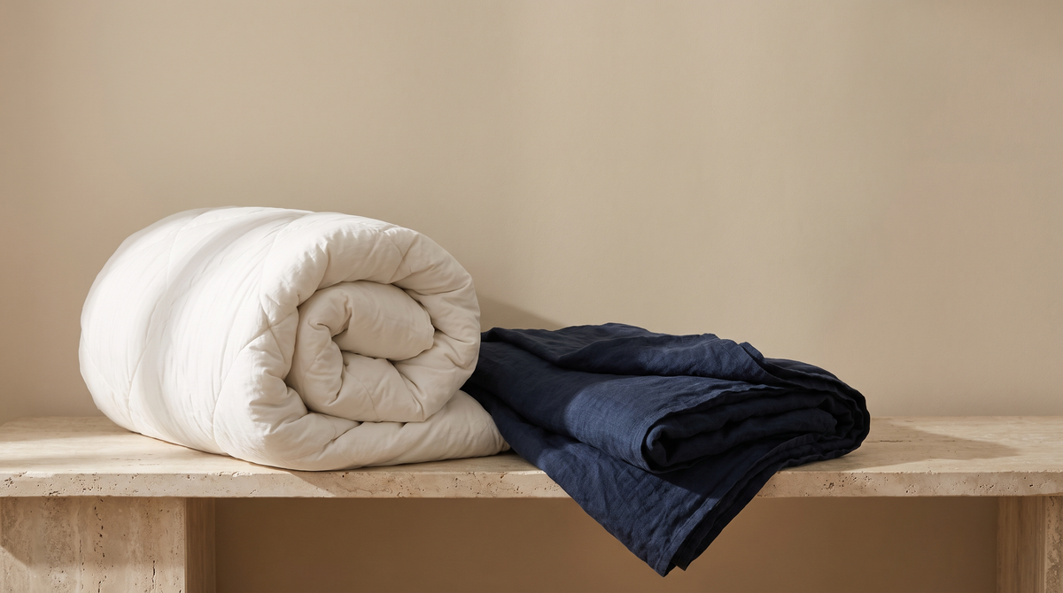 A thick white quilted duvet folded into a loose roll on a pale travertine ledge beside a folded deep navy linen throw in soft morning daylight