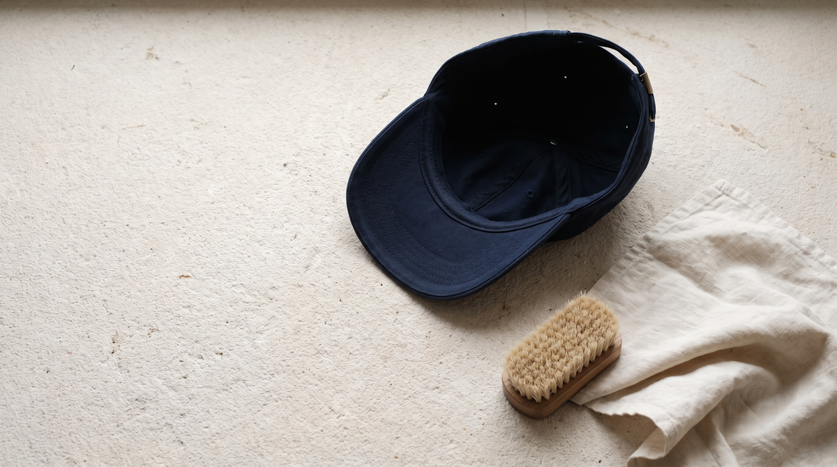 A plain deep navy cotton cap resting upside down on a pale limestone surface beside a soft bristle brush and a folded off-white linen cloth