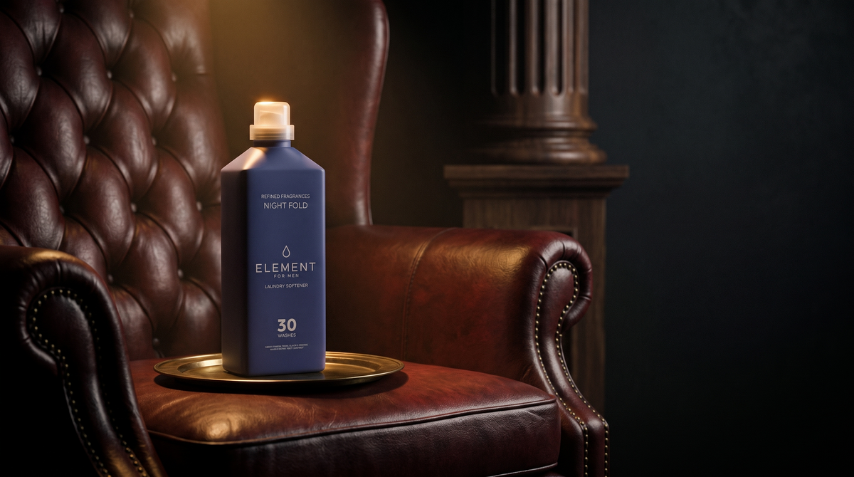 An Element for Men Night Fold fabric softener bottle standing on a brass tray on an oxblood leather wing chair, lit by a narrow warm spotlight