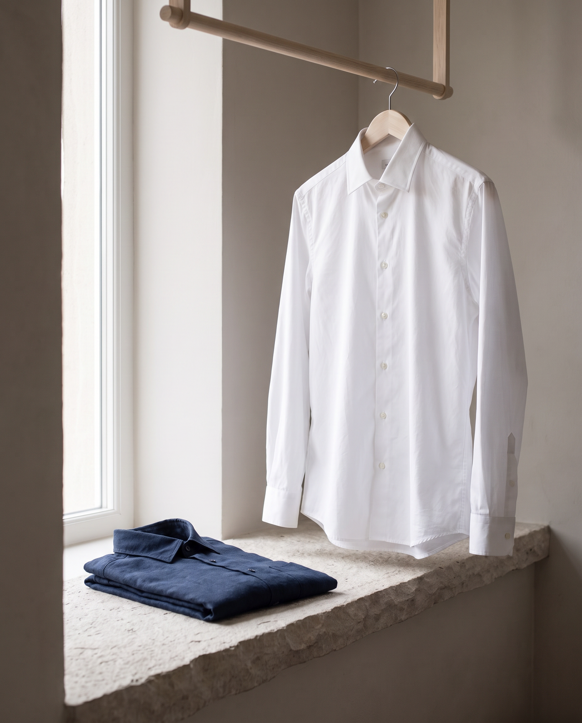 A crisp white shirt on a wooden hanger by a tall window, with a folded navy linen shirt on a pale limestone ledge